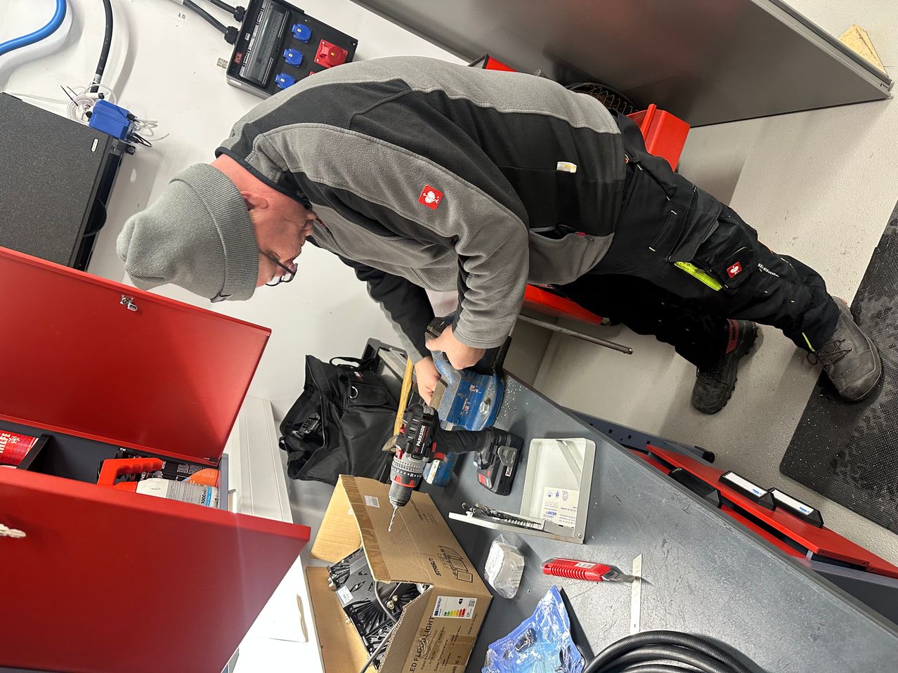 Ein Mann mit grauer Mütze und Brille biegt sich vor und benutzt eine Bohrmaschine auf einer Werkbank. Verschiedene Werkzeuge und Kisten sind auf der Werkbank, mit einem offenen roten Werkzeugkasten neben ihm.