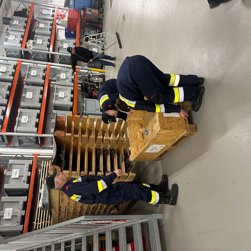 Mehrere Arbeiter in blauen Uniformen arbeiten in einem Lager. Zwei werden beim Knien und Untersuchen von Holzkisten gesehen, während ein weiterer den Boden fegt.