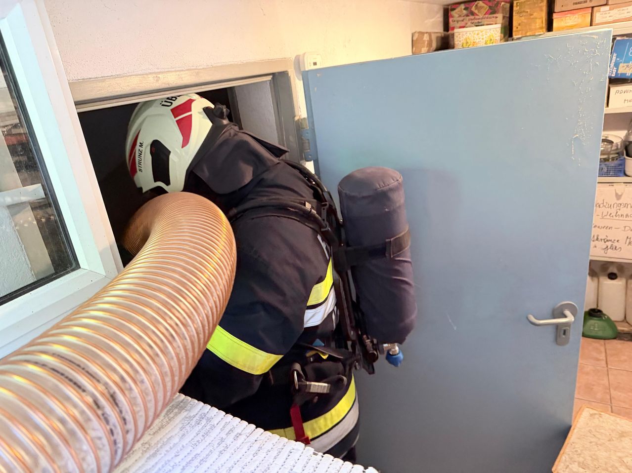 Ein Feuerwehrmann kriecht durch ein kleines Fenster und ist mit einem Atemschutzgerät ausgestattet. Er trägt einen weißen Helm und eine schwarze Uniform mit gelben reflektierenden Streifen.