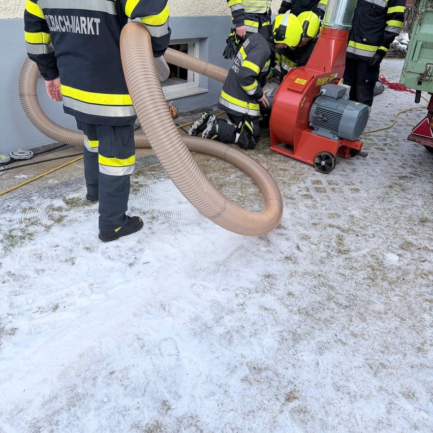 Feuerwehrleute benutzen einen roten Blaser, um Schnee von einem Gebäudeeingang zu entfernen. Ein Feuerwehrmann kniet, während ein anderer in der Nähe mit einem Schlauch steht.