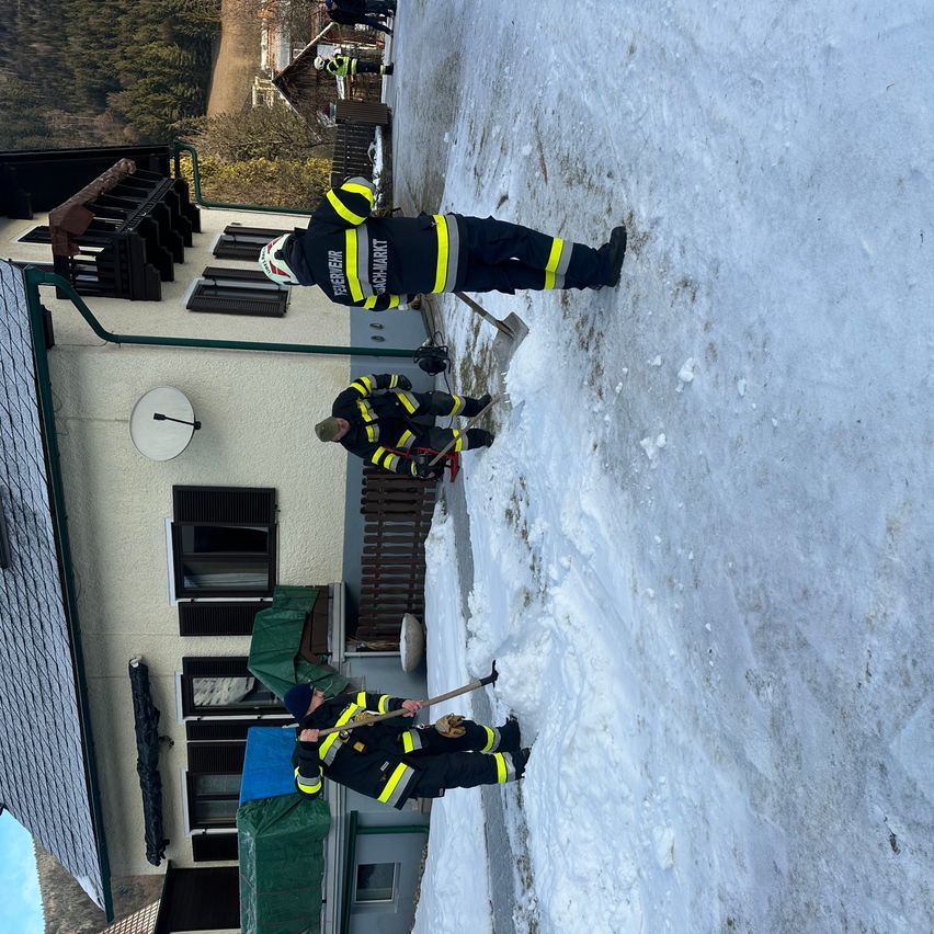 Drei Feuerwehrleute räumen Schnee von einer steilen Wand vor einem Gebäude. Sie tragen gelbe und schwarze Uniformen.