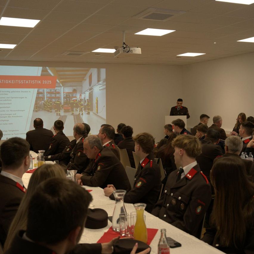 Eine Gruppe von Menschen in Uniformen sitzt in einem Raum mit einem Podium. Ein Mann steht am Podium und spricht die Zuhörer an. Eine Projektionsleinwand zeigt Informationen.