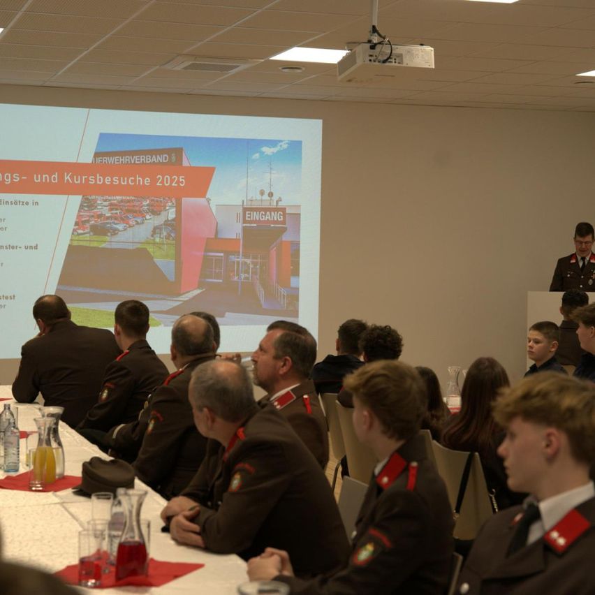 Eine Gruppe von Militärangehörigen in Uniform ist in einem Raum für eine Präsentation versammelt. Eine Projektionsleinwand zeigt eine Folie, und ein Mann in Uniform steht an einem Podium. Der Raum ist mit Stühlen, Tischen und Erfrischungen gefüllt.