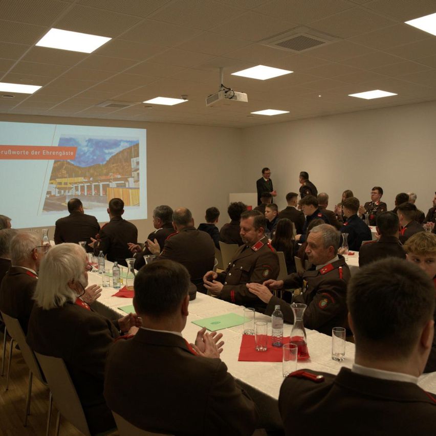 Eine Gruppe uniformierter Personen sitzt an Tischen in einem Raum und hört einer Präsentation auf einer Projektionsleinwand zu. Der Moderator steht an einem Podium, und einige Teilnehmer tragen Brillen. Die Umgebung ist feierlich, mit einer weißen Wand und Deckenleuchten.