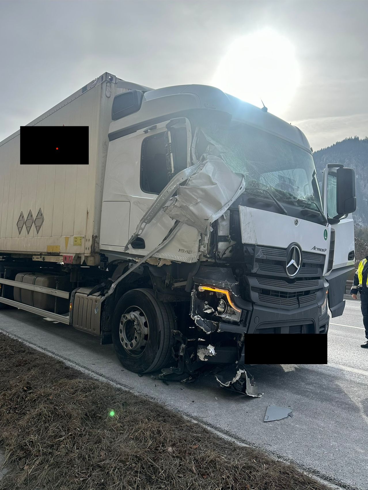 A white Mercedes-Benz truck has suffered significant front-end damage, with the bumper and hood bent and the windshield shattered. There are two black-covered areas on the image, possibly to obscure sensitive information.