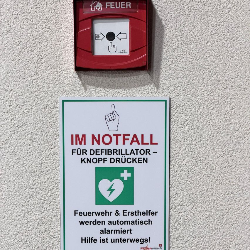Eine rote Feueralarmbox ist an einer weißen Wand montiert. Ein weißes Schild darunter weist darauf hin, in Notfällen einen Knopf für einen Defibrillator zu drücken.