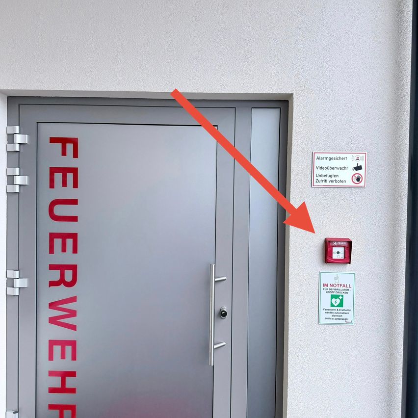 Eine graue Tür mit dem Wort 'Feuerwehr' in Rot. Ein Pfeil zeigt auf einen Feueralarm, ein Schild für Videoüberwachung und einen Defibrillator.