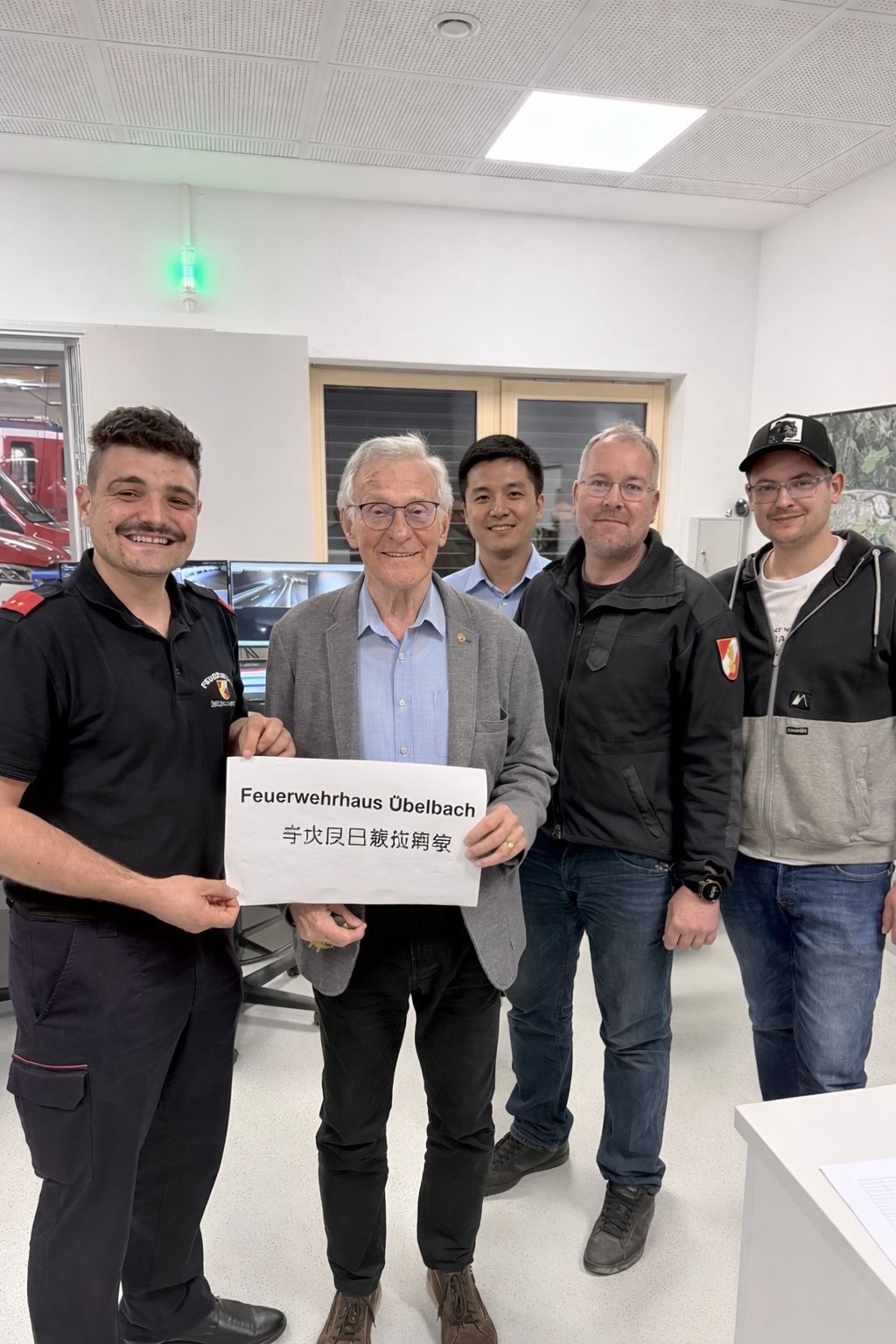 Fünf Männer stehen in einem Raum, von denen einer ein Schild mit der Aufschrift 'Feuerwehrhaus Ubelbach' hält. Sie lächeln und scheinen für ein Foto zu posieren.