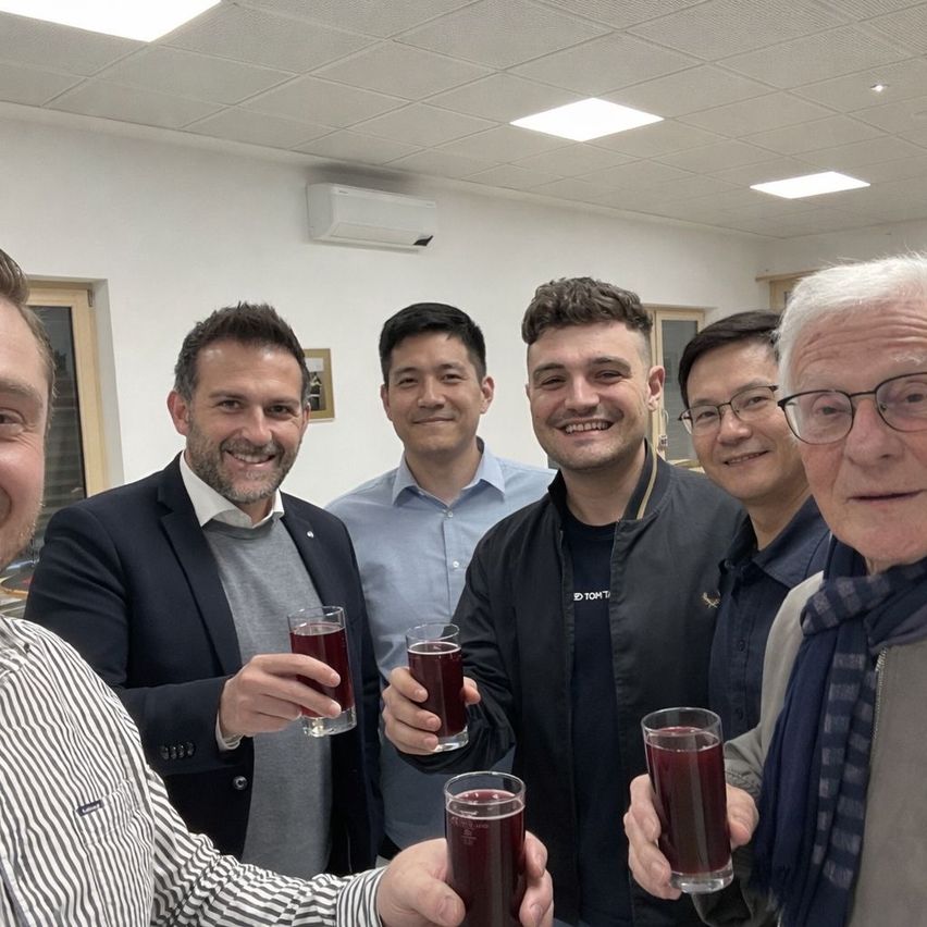 Eine Gruppe von Männern, alle lächelnd und Gläser mit roter Flüssigkeit in der Hand, stehen gemeinsam in einem Raum. Sie scheinen zu feiern.