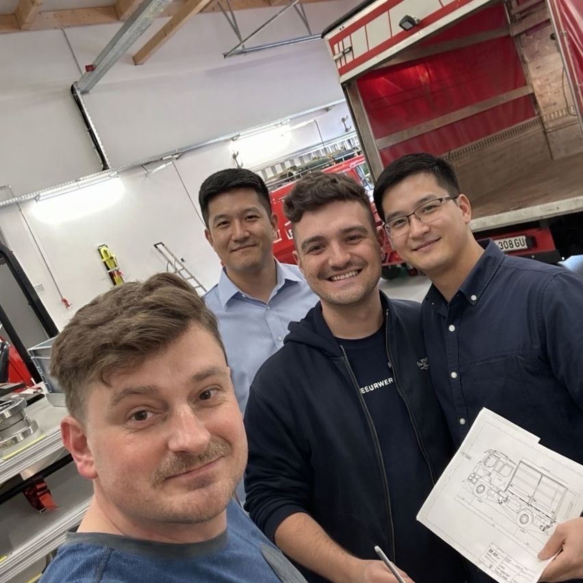 Drei Männer stehen in einer Garage mit einem Feuerwehrauto dahinter. Einer hält eine Blaupause, und alle lächeln für ein Foto.