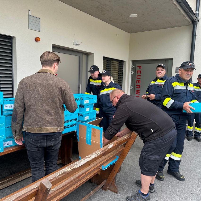 A group of firefighters are sorting through boxes in a warehouse. A man in a brown jacket and jeans stands near a table.