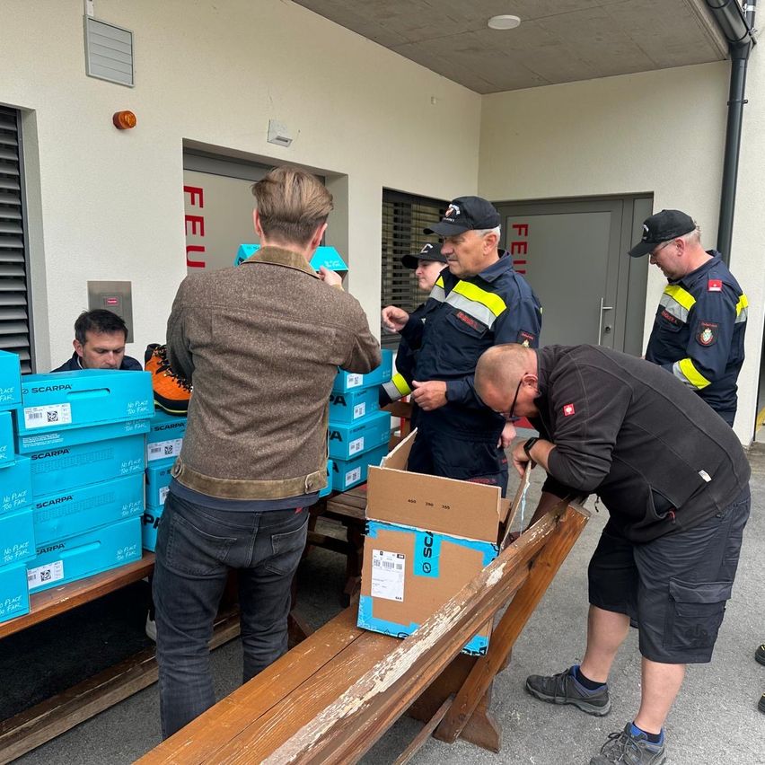 A group of men in uniforms are sorting through a box of Scarpa shoes. They are standing around a wooden table with boxes of Scarpa shoes on it.