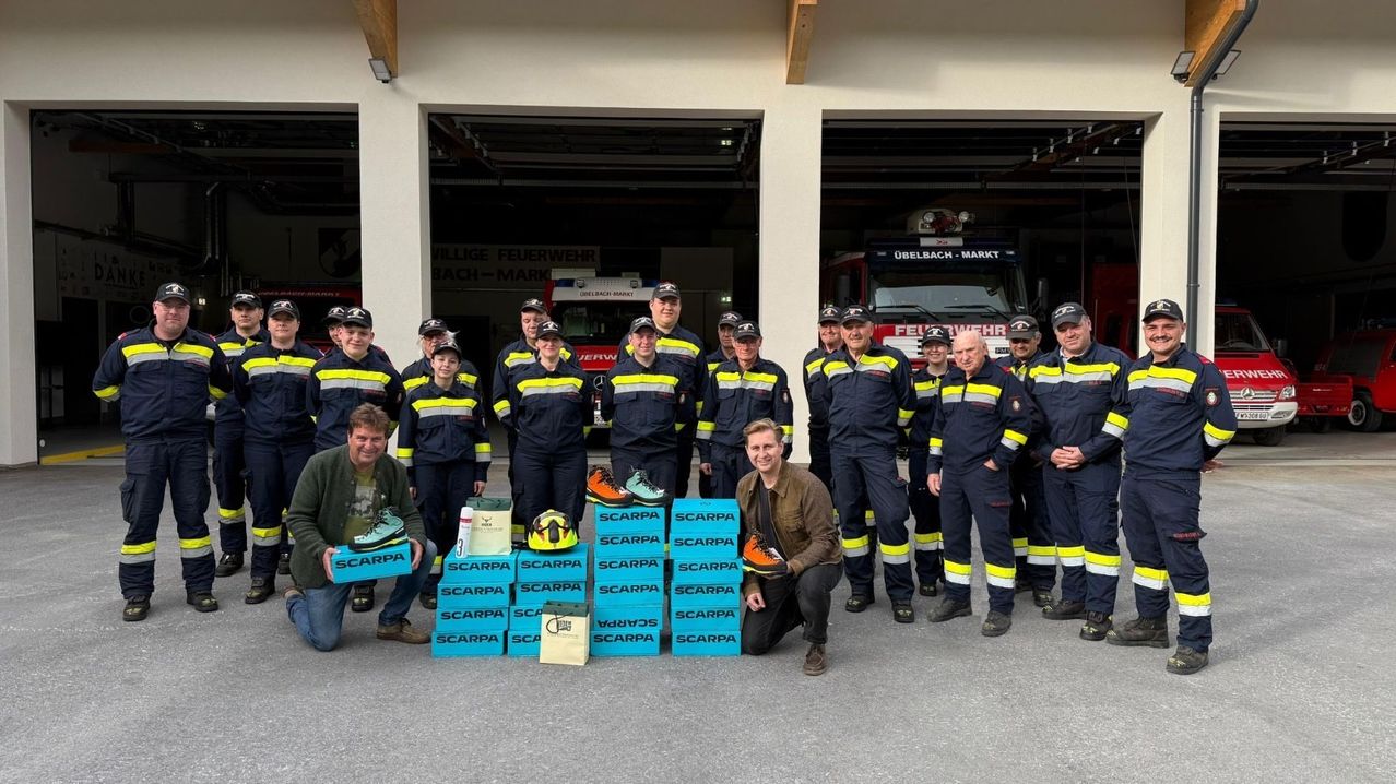 A group of firefighters poses with boxes of Scarpa shoes, helmets, and bags in front of a fire station.