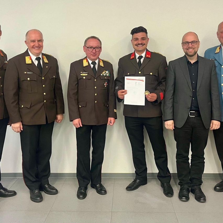 Five men in uniform pose for a photo. One holds a certificate, smiling. The others wear ties and uniforms. They stand in front of a white wall.