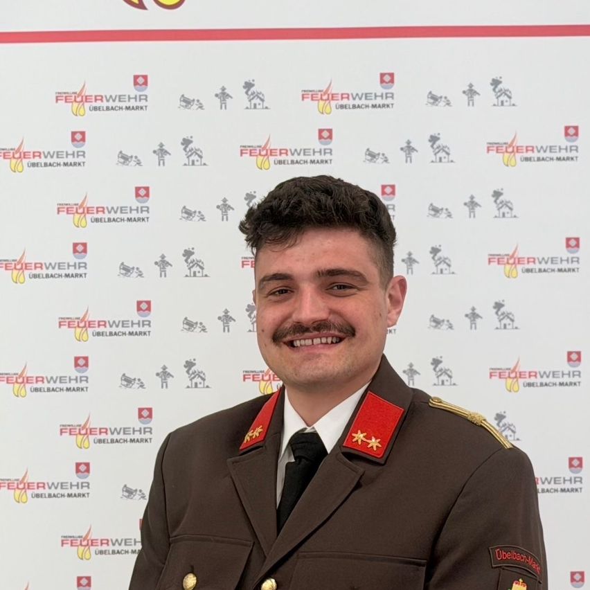A man in a military uniform with a smiling face is standing in front of a wall with logos. He has a mustache and is looking straight ahead.
