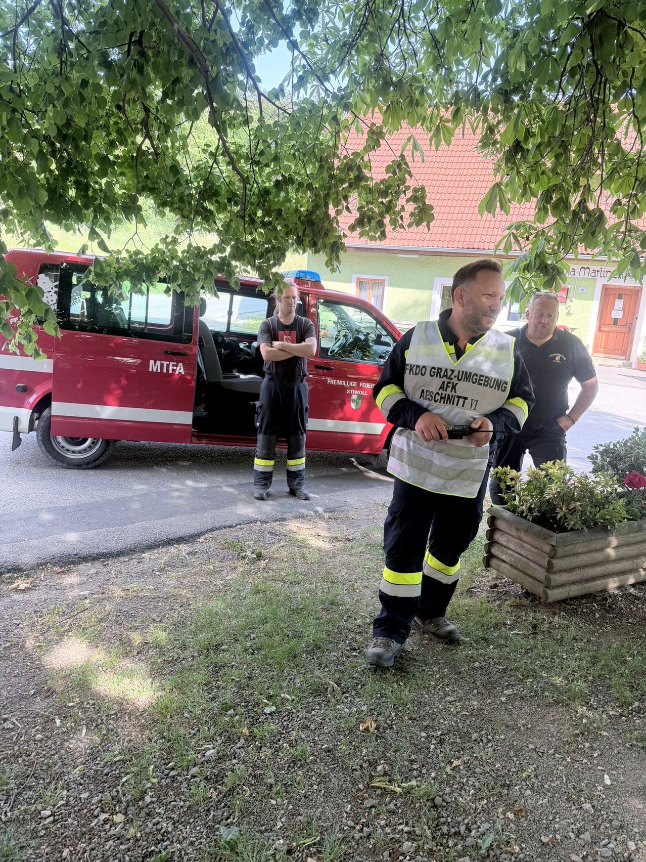 Drei Männer in Uniformen stehen vor einem roten Feuerwehrauto mit offener Tür vor einem Haus.