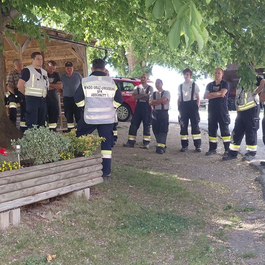 Eine Gruppe von Feuerwehrleuten steht in einer Reihe unter einem Baum. Einige Leute stehen dahinter.