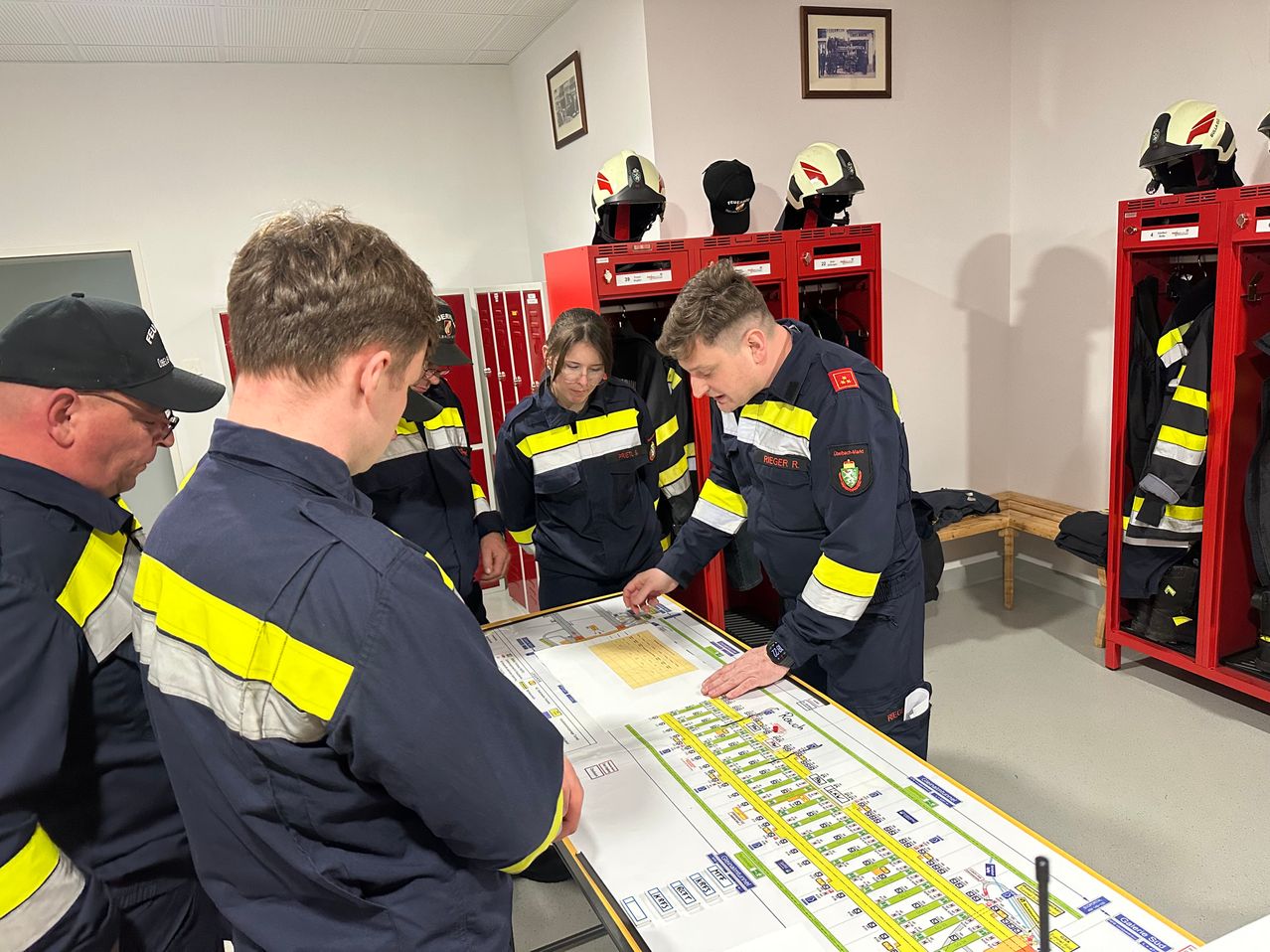 Eine Gruppe von Feuerwehrleuten ist um eine Karte in einem Raum mit roten Schließfächern und Helmen darüber versammelt.