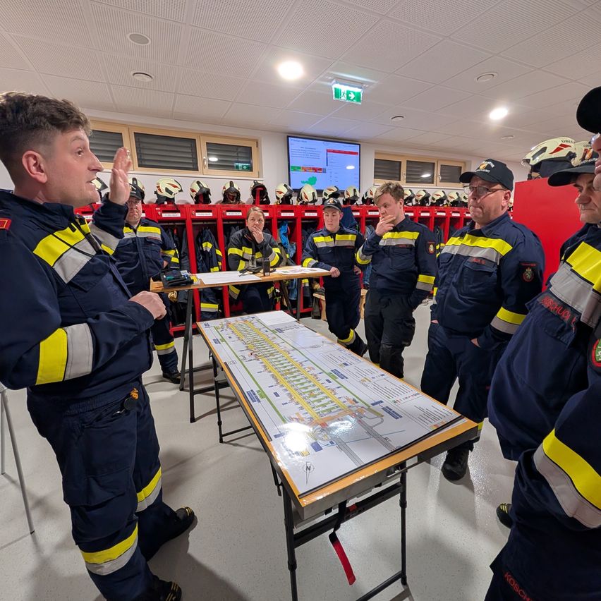 Feuerwehrleute versammeln sich in einem Raum und diskutieren über eine Karte auf einem Tisch. Sie tragen blaue Uniformen mit gelben und roten Streifen.