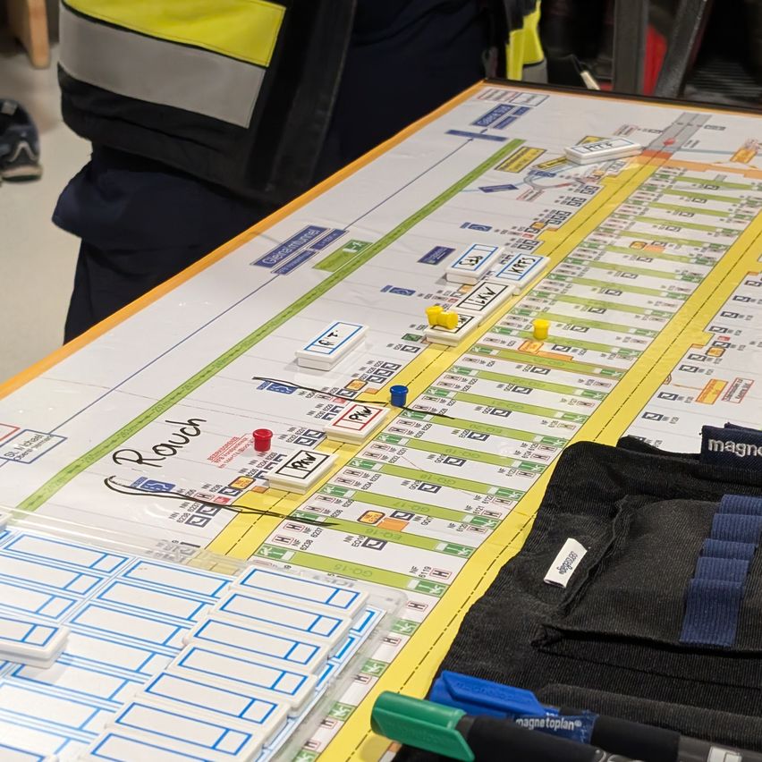 Ein Brett mit verschiedenen farbigen Markierungen und Etiketten steht auf einem Tisch. Eine Person in einer reflektierenden Weste steht dahinter. Das Brett zeigt ein kartenähnliches Layout mit Gittern und Symbolen.