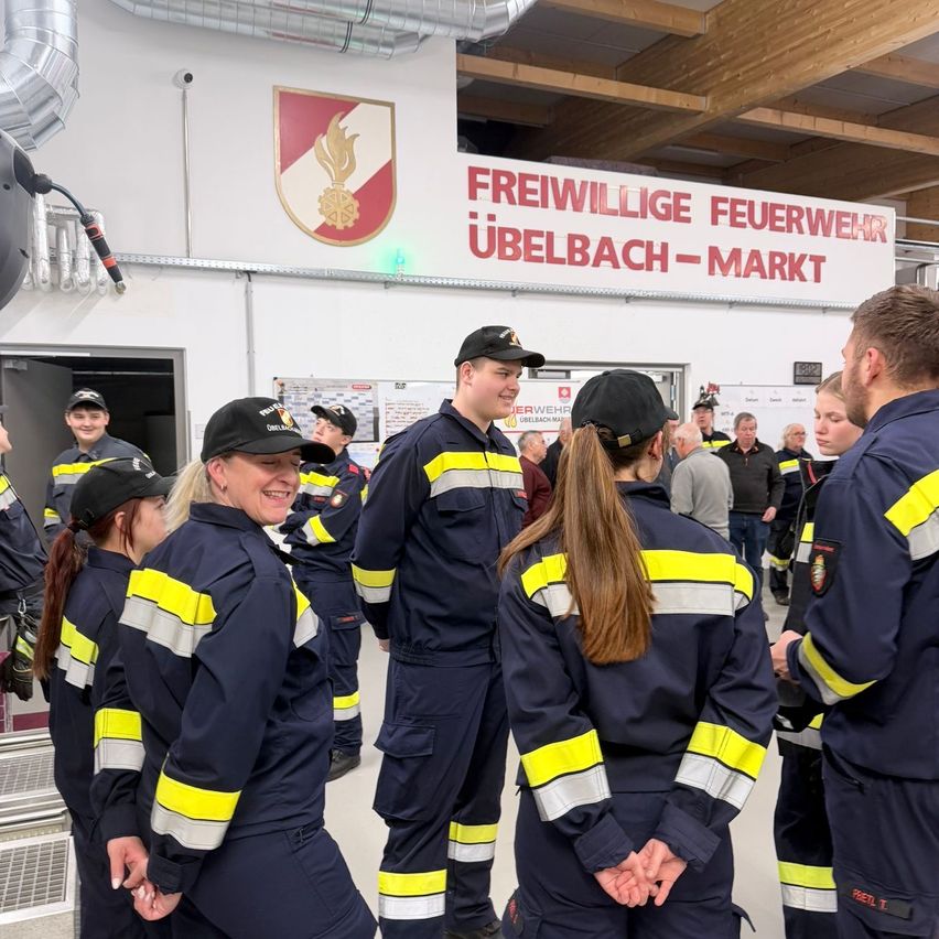 Freiwillige Feuerwehrleute versammeln sich in der Feuerwache Ubelbach-Markt, lächelnd und plaudernd, in ihren Uniformen mit reflektierenden Streifen. Der Eingang der Wache steht offen, und ein Schild mit Feuersymbol ziert die Wand.