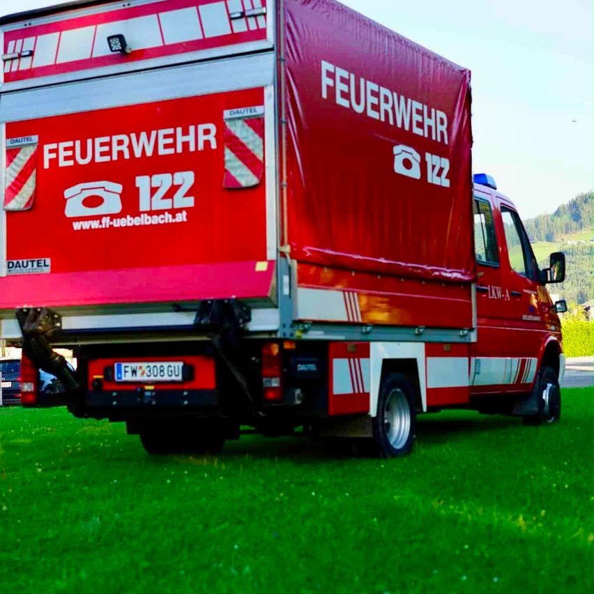A red fire department truck with the number 122 and the website www.ff-uebebach.at on the side, parked on a grassy field.