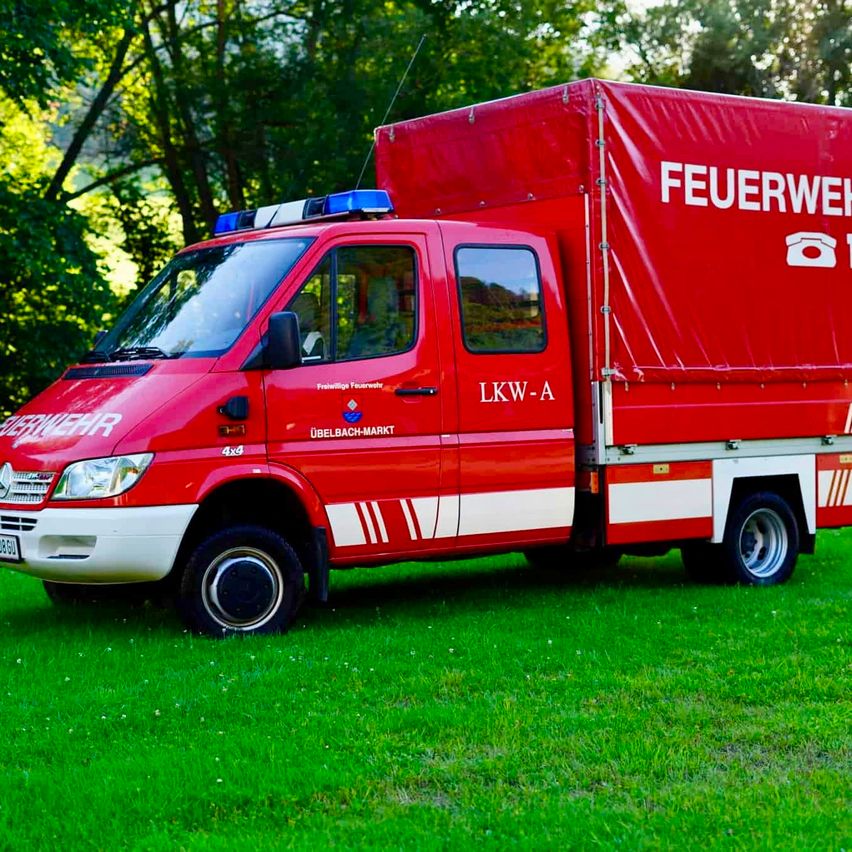 A red fire truck with the text 'Feuerwehr' on the side is parked on a green lawn. The truck has a blue light on top and a white stripe on the side.