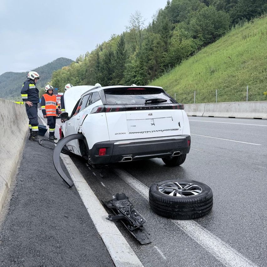 Ein weißer SUV ist am Straßenrand geparkt, wobei die Heckpartie beschädigt ist. Zwei Personen in Sicherheitsuniformen stehen in der Nähe und inspizieren möglicherweise das Fahrzeug. Ein Reifen und Felge liegen auf der Straße, und eine Barriere befindet sich an der Seite.