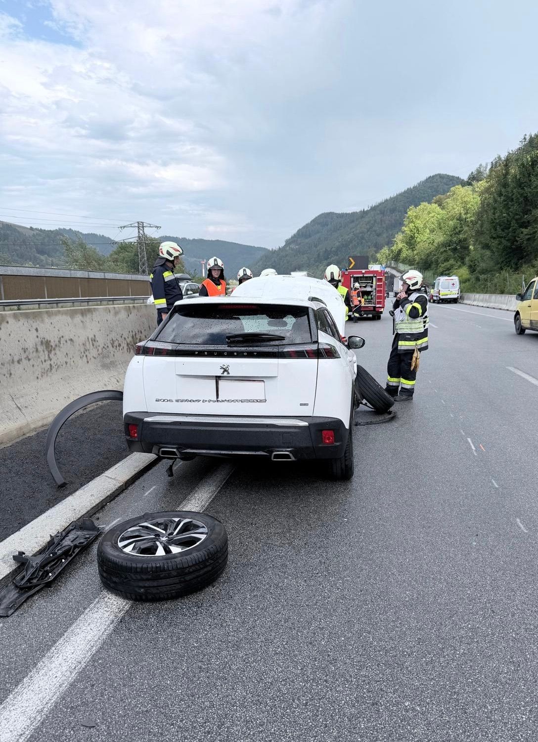 Rettungskräfte kümmern sich um einen weißen SUV am Straßenrand, mit einem nahegelegenen platten Reifen und einem Feuerwehrwagen im Hintergrund.