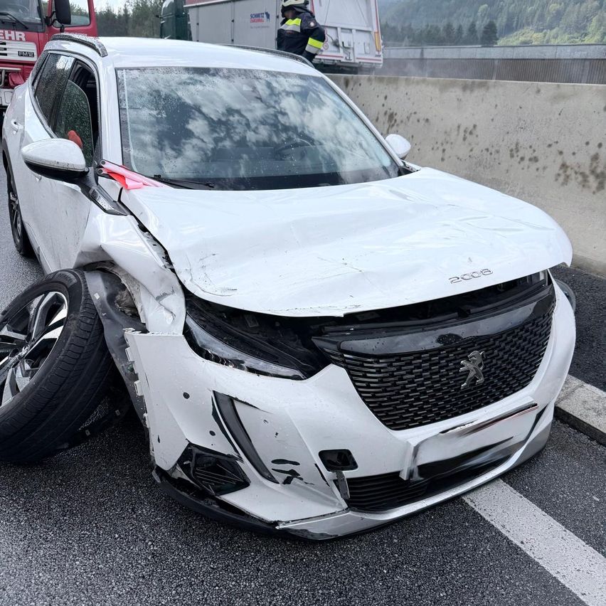 Ein weißer Peugeot SUV mit schweren Schäden an der Front steht am Straßenrand, mit einem platten Reifen und einer Person in einer reflektierenden Weste in der Nähe.