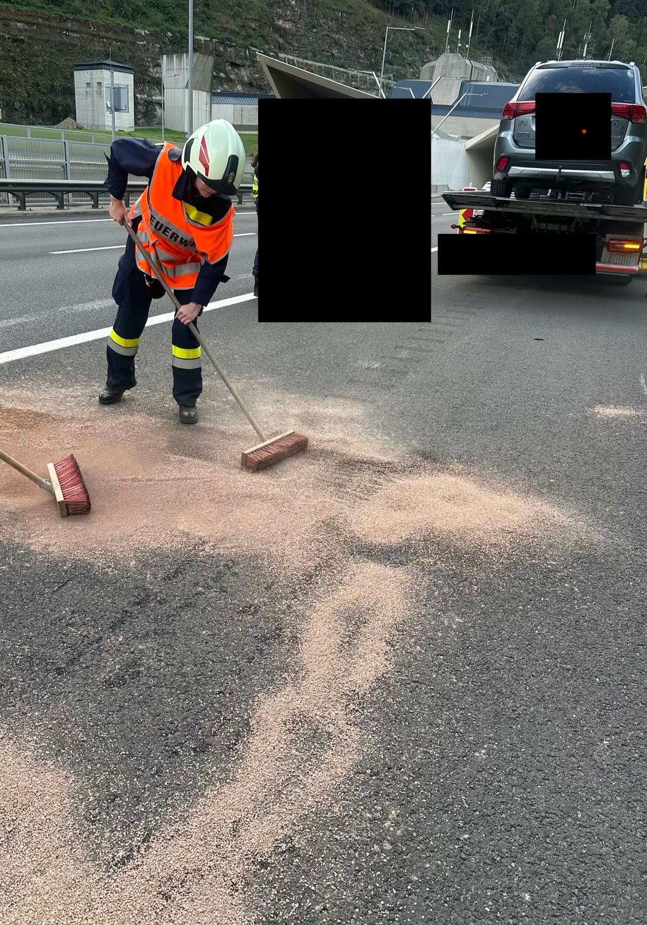 Ein Arbeiter in einer reflektierenden Weste und Helm fegt auf einer Straße Schmutz auf. Ein Abschleppwagen ist im Hintergrund zu sehen.
