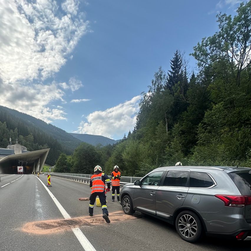 Zwei Arbeiter in orangen Westen säubern eine Verschüttung auf einer Autobahn, in der Nähe ist ein Auto geparkt.