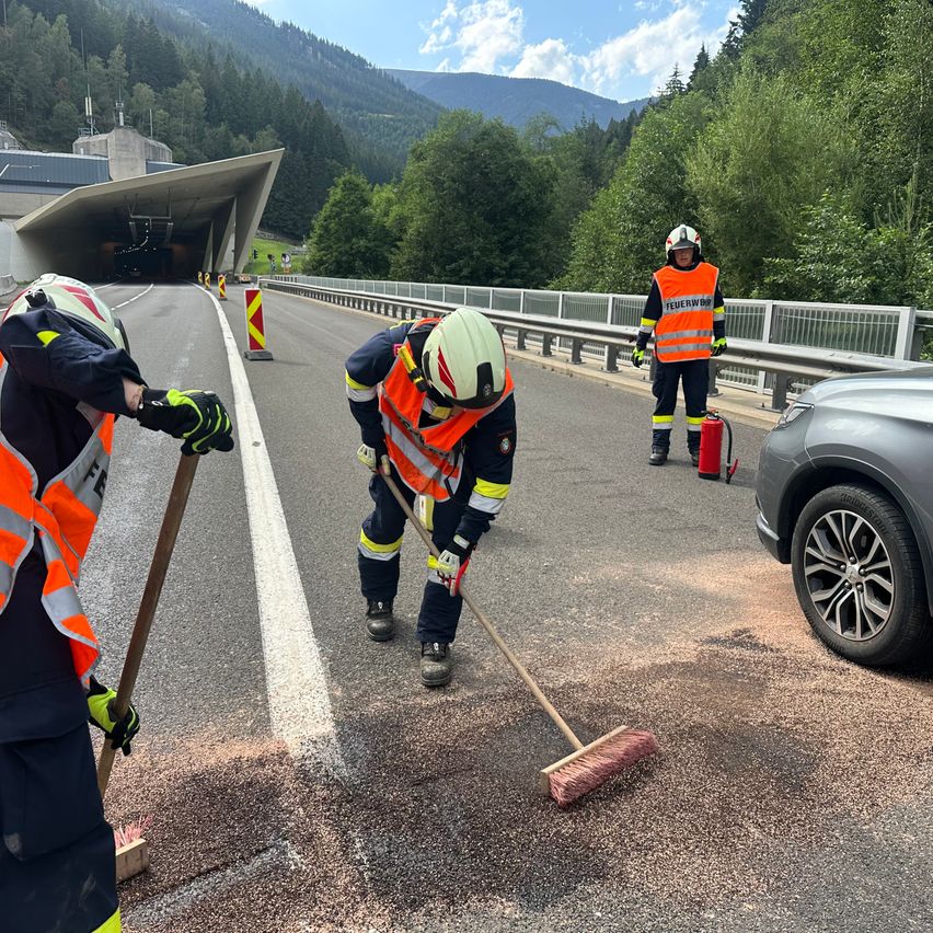 Drei Arbeiter in Sicherheitsausrüstung reinigen die Straße, einer mit einem Besen, ein anderer mit einem Feuerlöscher in der Nähe, und ein Auto parkt an der Seite.
