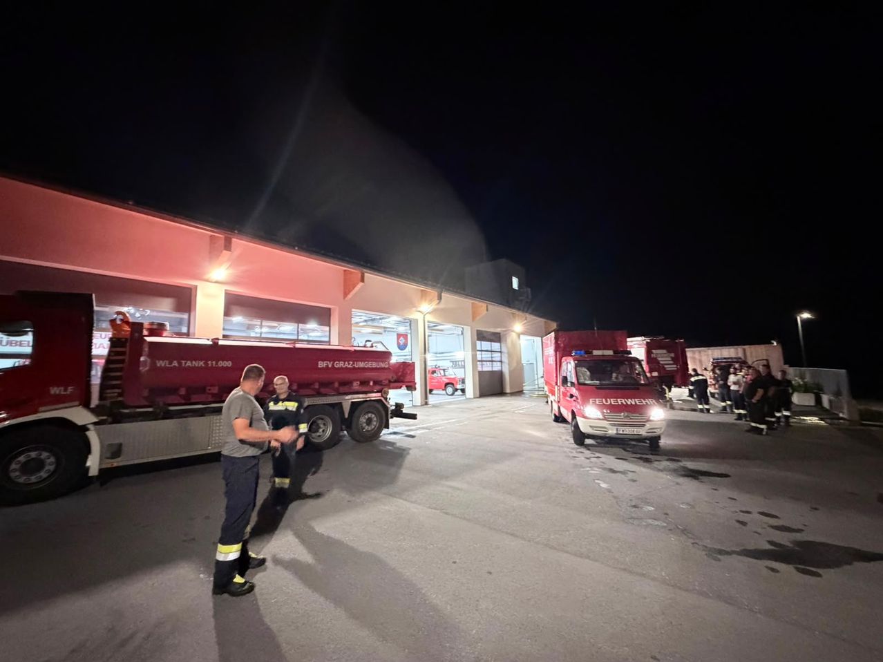 Firefighters are gathered around a fire truck at night in front of a fire station. The fire truck has the words 'Feuerwehr' on it.