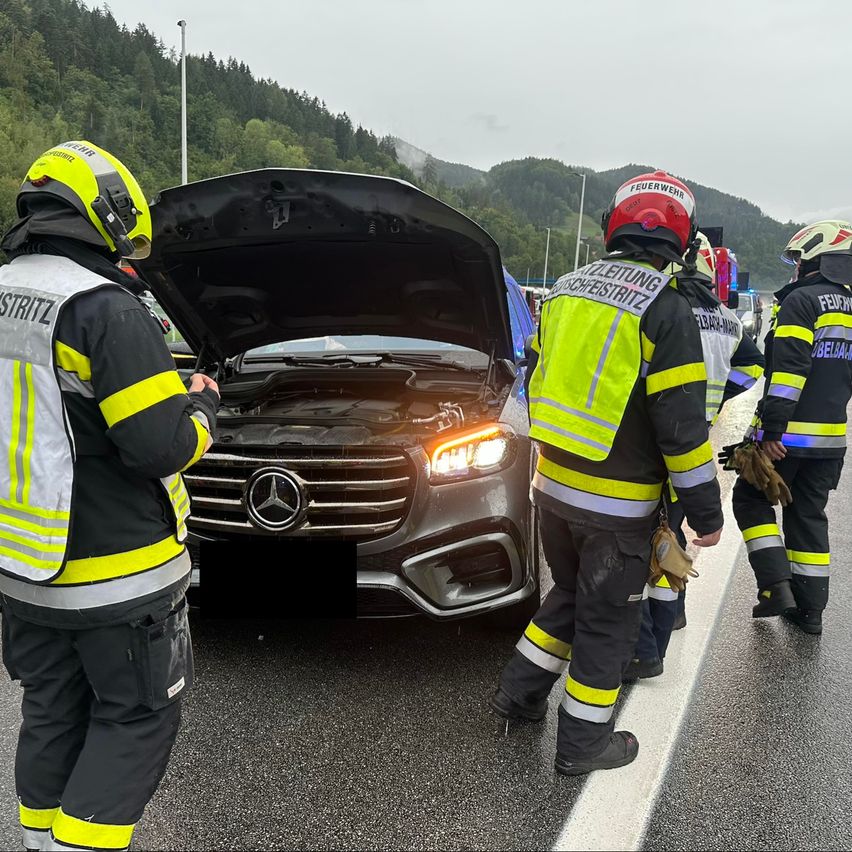 Feuerwehrleute in Schutzkleidung inspizieren einen Mercedes-Benz mit offenem Motorhaube am Straßenrand, mit Bergen im Hintergrund.