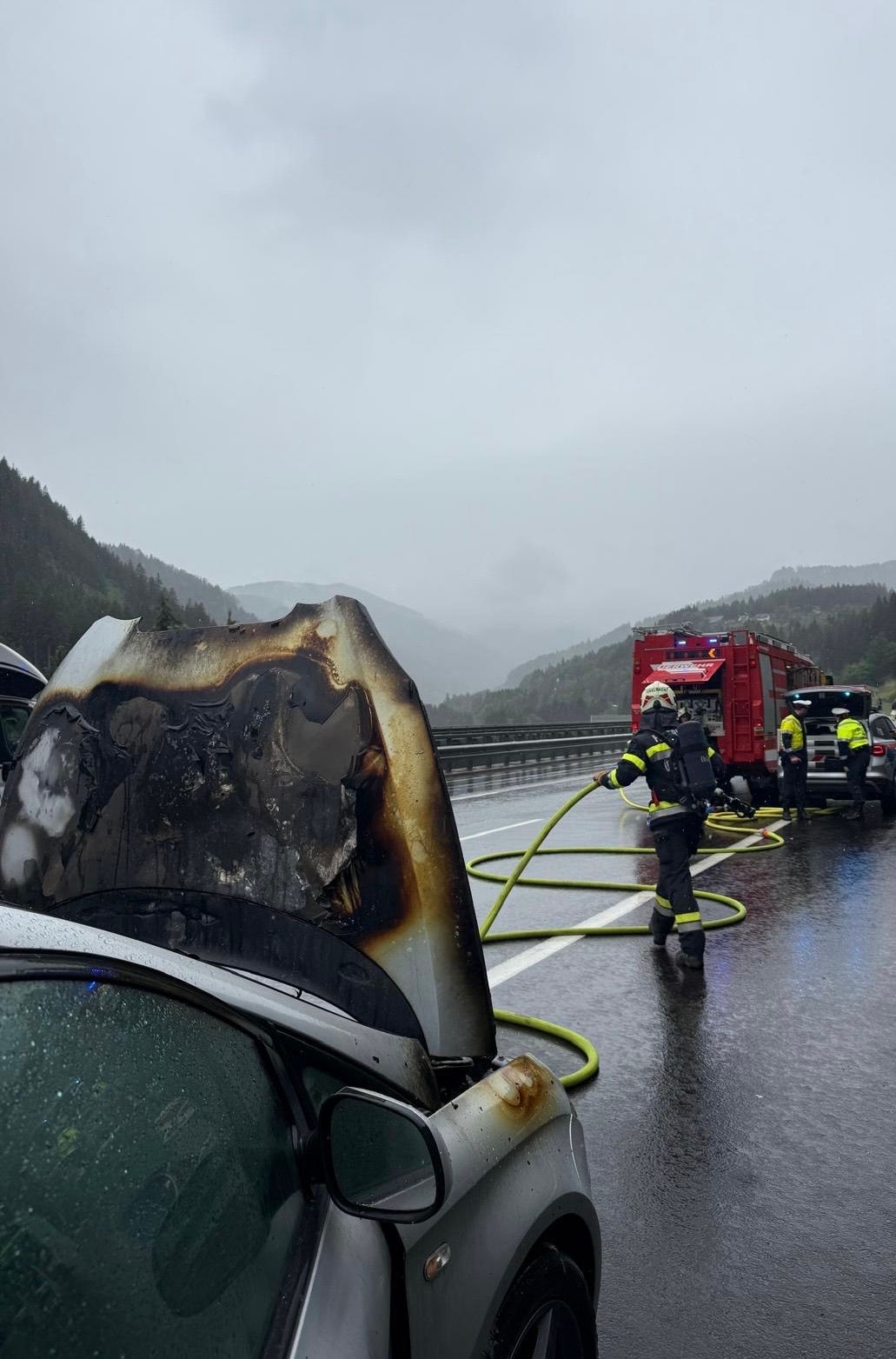 Firefighters in rain gear extinguishing a burned car on a highway. The car's hood is open, revealing a damaged engine. A fire truck is parked nearby.