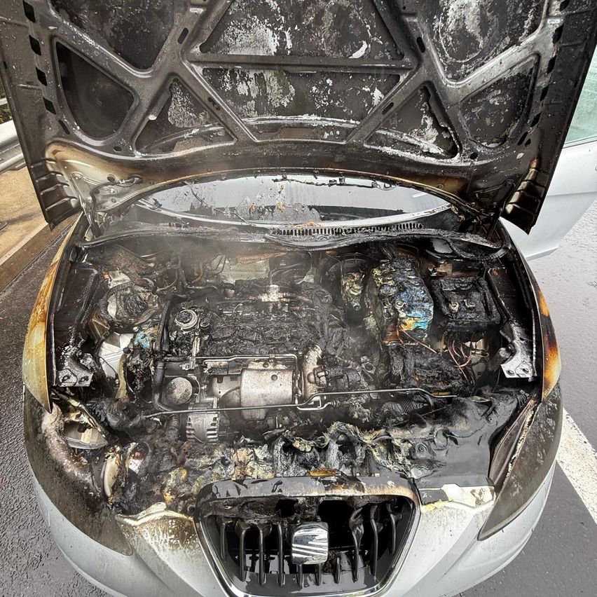 A car's engine compartment is open, revealing a charred and damaged engine. Smoke is coming from the engine area, and the hood is lifted. The front bumper is also damaged.