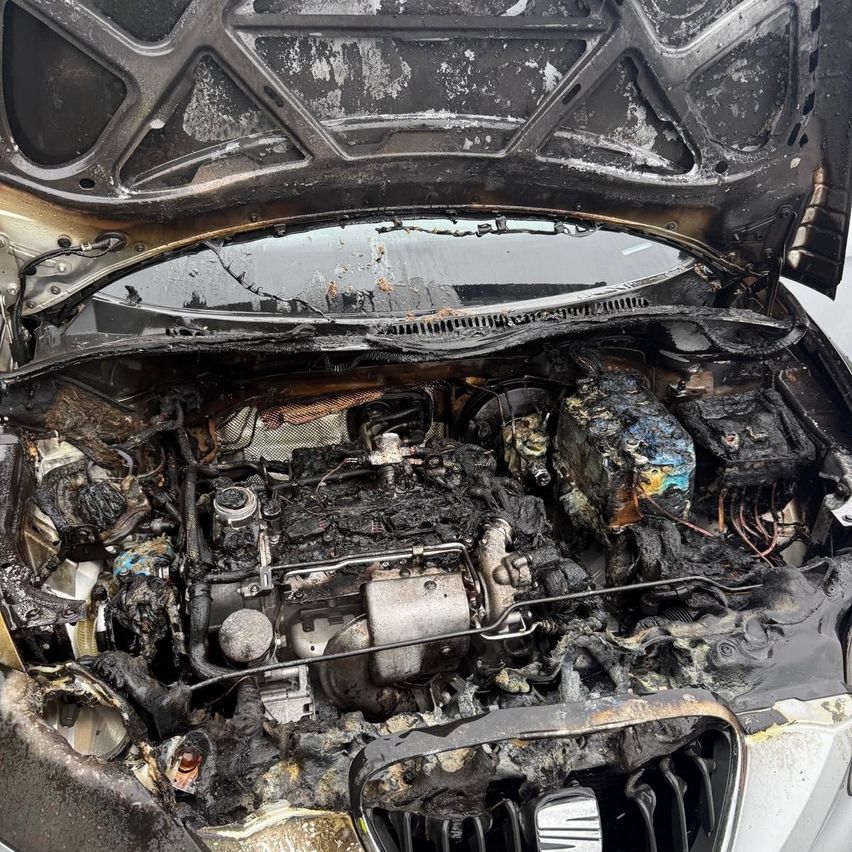 The engine compartment of a car is open, showing the burned and damaged engine with black soot and melted parts.
