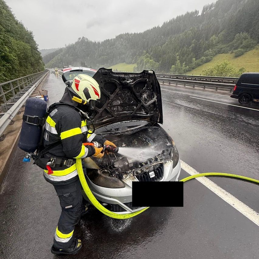 A firefighter is using a hose to extinguish a car fire on a wet highway with mountains in the background.