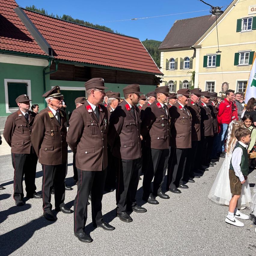 Military officers in brown uniforms stand in a line outdoors, with a group of people behind them. A young child in a white dress and a young boy in a green vest are standing in front of the officers. Buildings with red roofs and yellow walls are behind them.