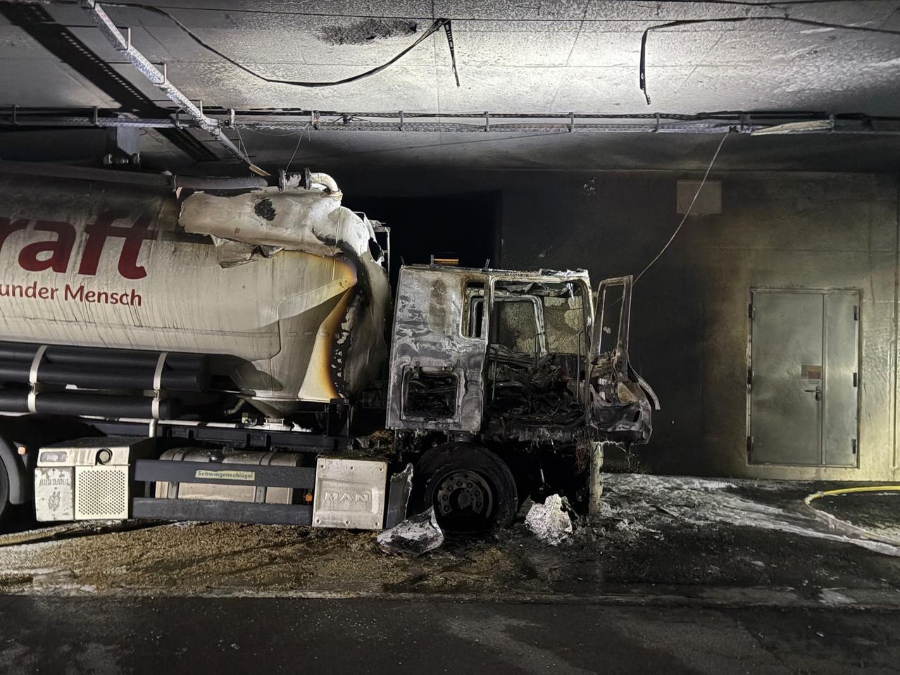 A severely burnt and damaged truck, with its front crushed and charred, rests in a dark, enclosed space. The ceiling shows signs of soot and damage.