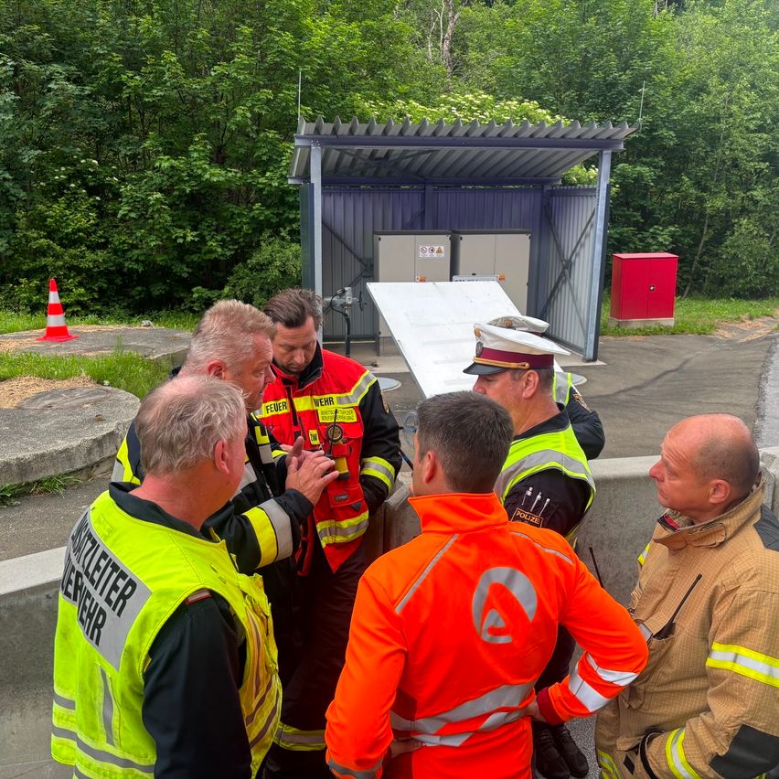 A group of emergency workers in safety gear converses near a small shelter, with a traffic cone and greenery in the background.