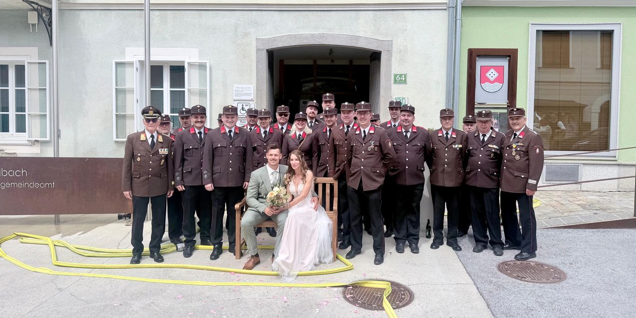 A group of uniformed individuals stand in front of a building with a bride and groom sitting on a bench. The bride holds a bouquet and the groom wears a suit.