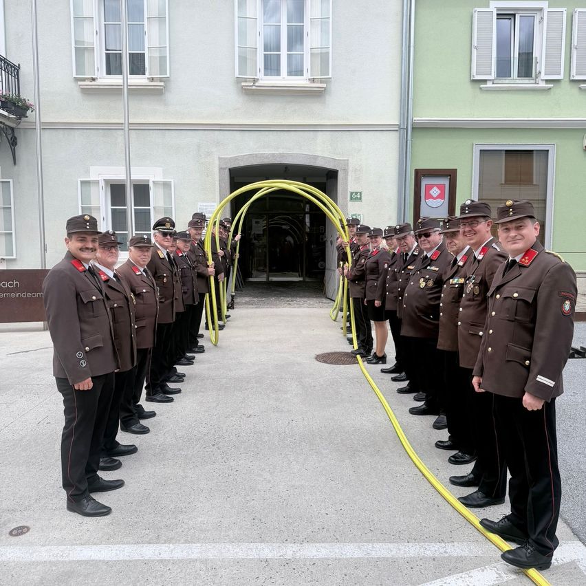 A line of military officers in uniform stands before a building with a yellow pipe. The building has white windows and a sign with a logo.