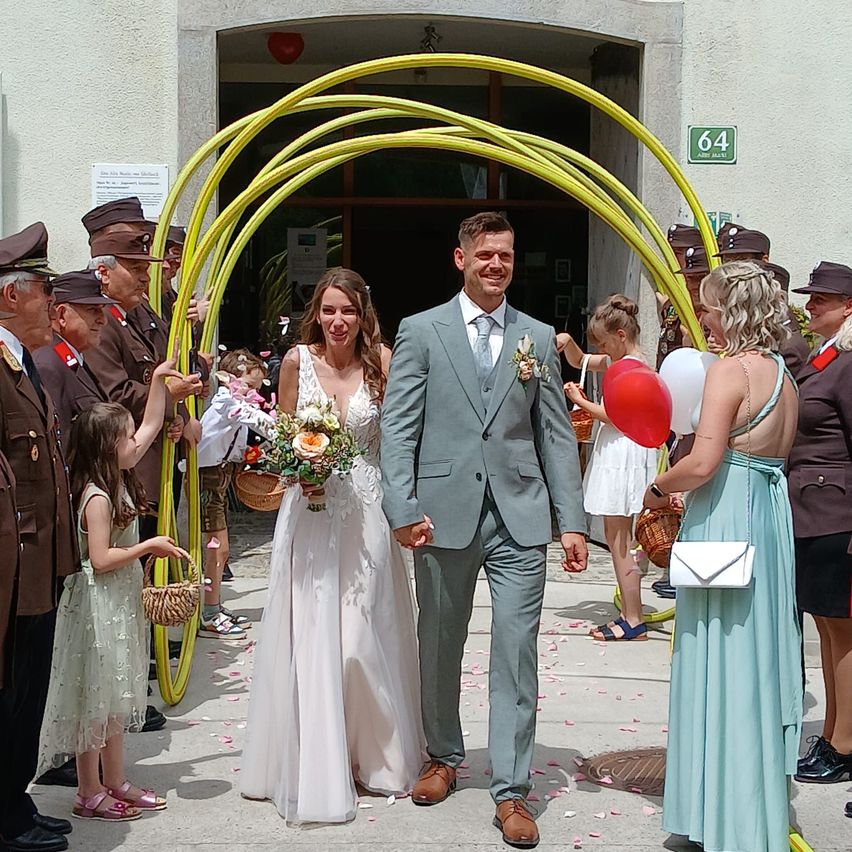 A bride and groom walk through an archway of yellow ribbons, surrounded by children and adults, some in uniforms. The bride holds a bouquet, the groom a bouquet of flowers. Behind them, people hold balloons and handbags.