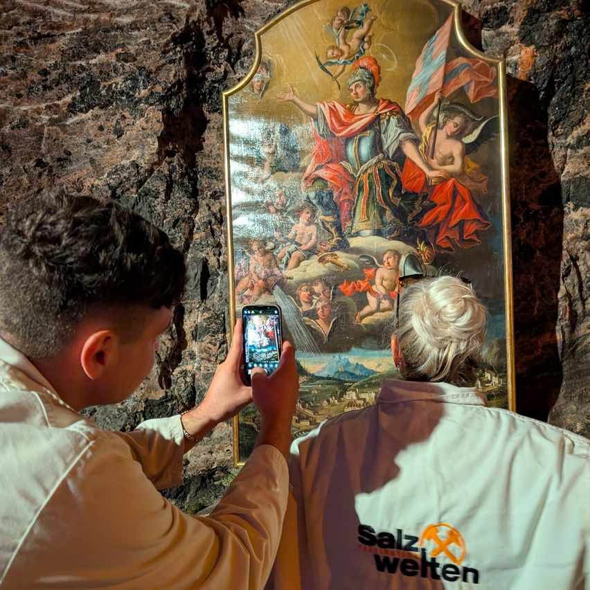 Zwei Personen in weißen Jacken stehen vor einem großen Gemälde an einer Steinwand. Einer macht ein Foto mit seinem Handy.