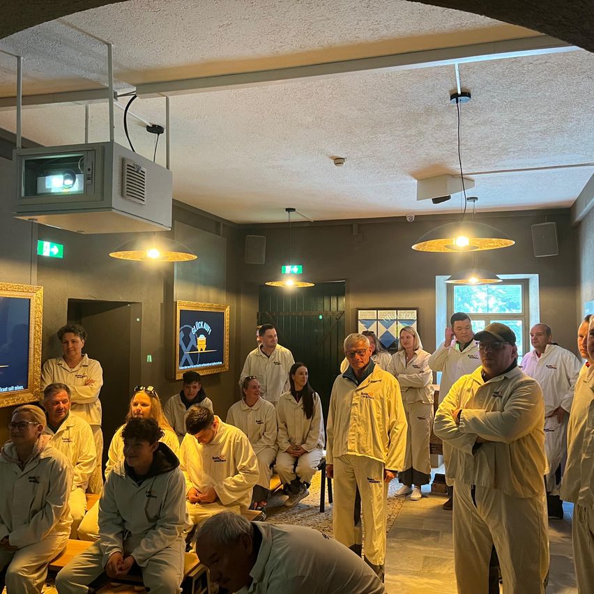 Eine Gruppe von Menschen in weißen Laborkittel steht in einem Raum. Einige sitzen auf Bänken, während andere stehen. Der Raum hat graue Wände und ist mit Deckenleuchten beleuchtet.