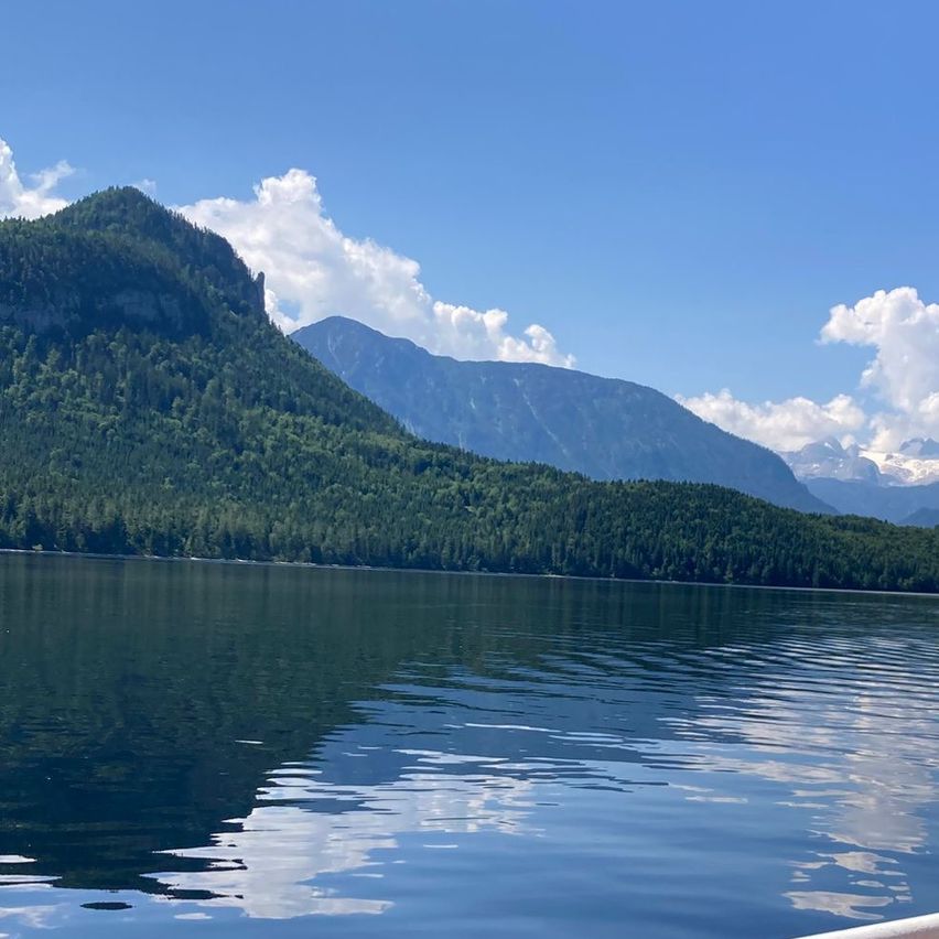 Ein ruhiger See mit Spiegelung des blauen Himmels und der Berge, umgeben von üppigen grünen Bäumen und Hügeln unter einem teilweise bewölkten Himmel.