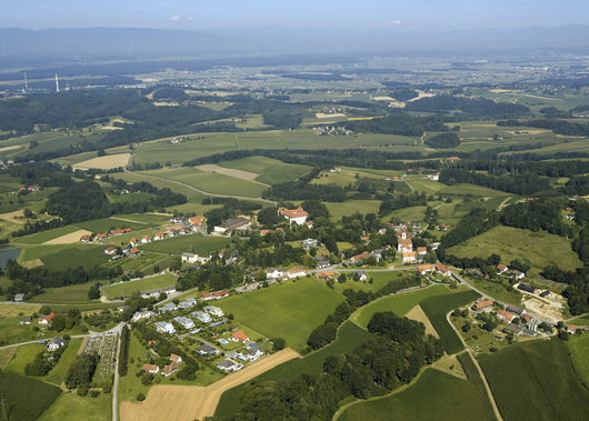 Luftaufnahme eines ländlichen Dorfes mit verstreuten Häusern, üppigen grünen Feldern und Bäumen, umgeben von Bergen in der Ferne.