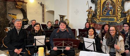 Eine Gruppe von Musikern versammelt sich in einer Kirche und spielt verschiedene Instrumente. Sie sitzen im Halbkreis mit Notenständern vor sich. Dahinter steht eine Kerze, und eine Statue eines Engels ist über dem Altar.